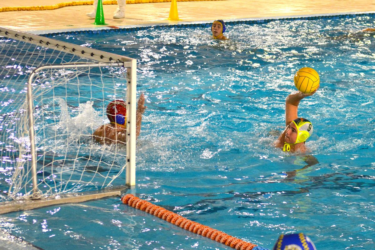 Jugador de waterpolo con gorro amarillo lanza el balón hacia la portería mientras el portero intenta detenerlo.