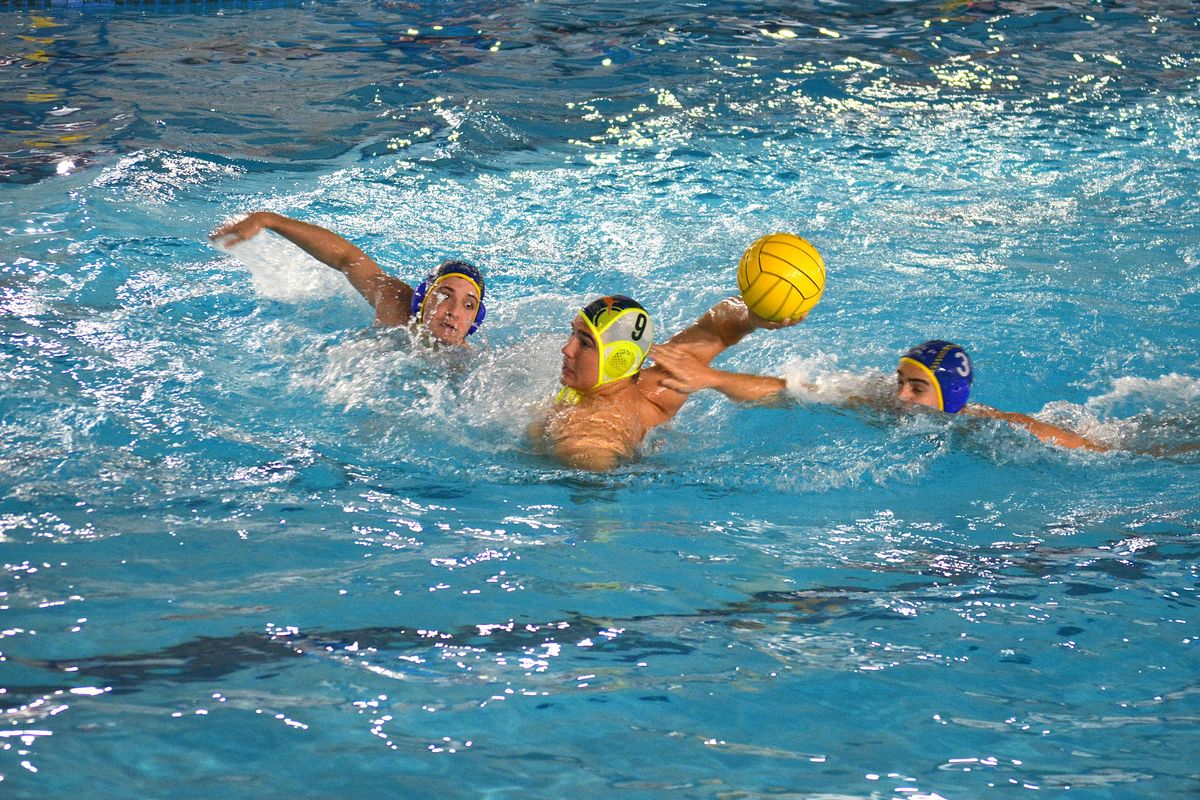 Jugador de waterpolo progresa con el balón amarillo mientras dos rivales intentan defender la jugada.