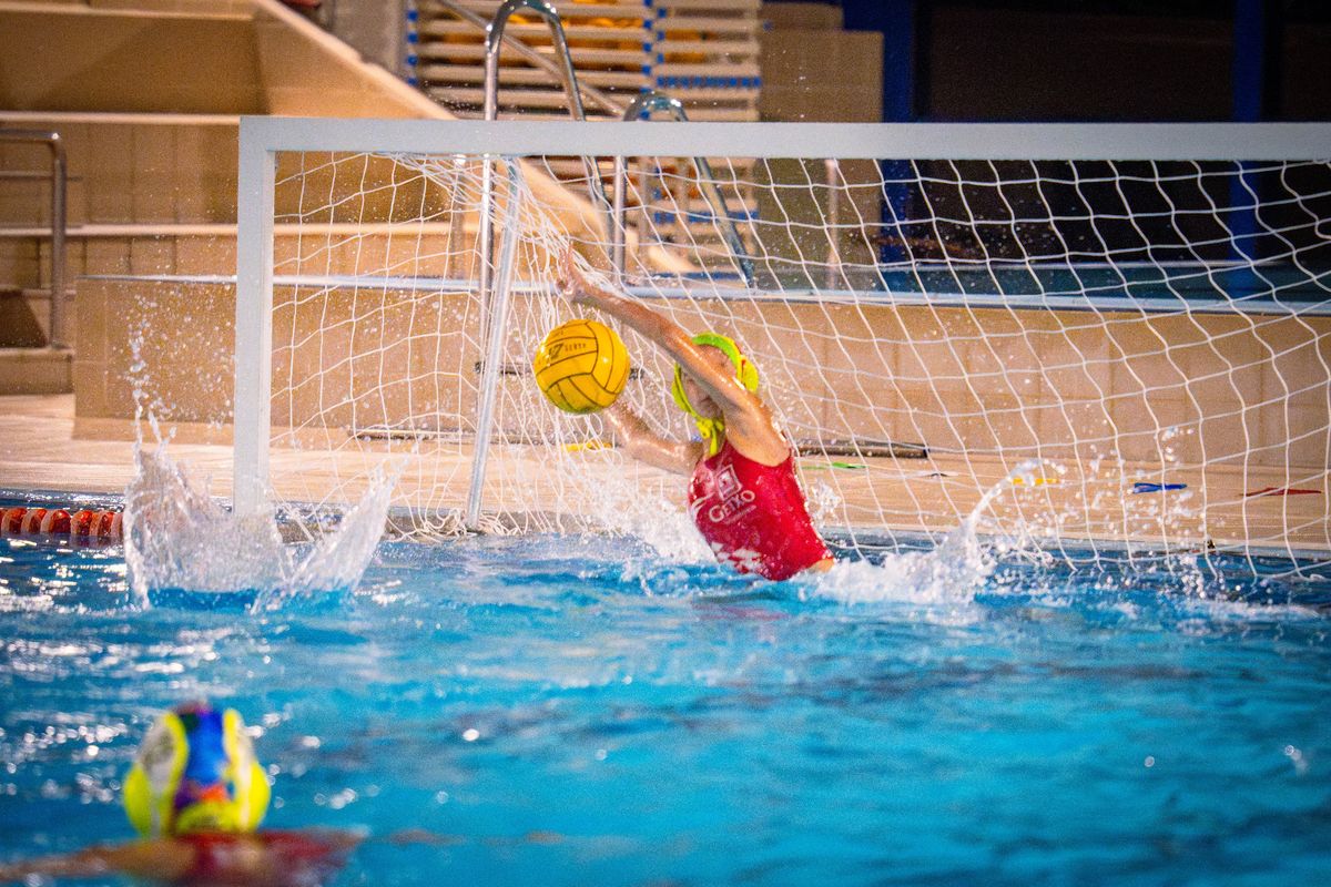 Portera de waterpolo con bañador rojo intenta detener un balón amarillo frente a la portería.