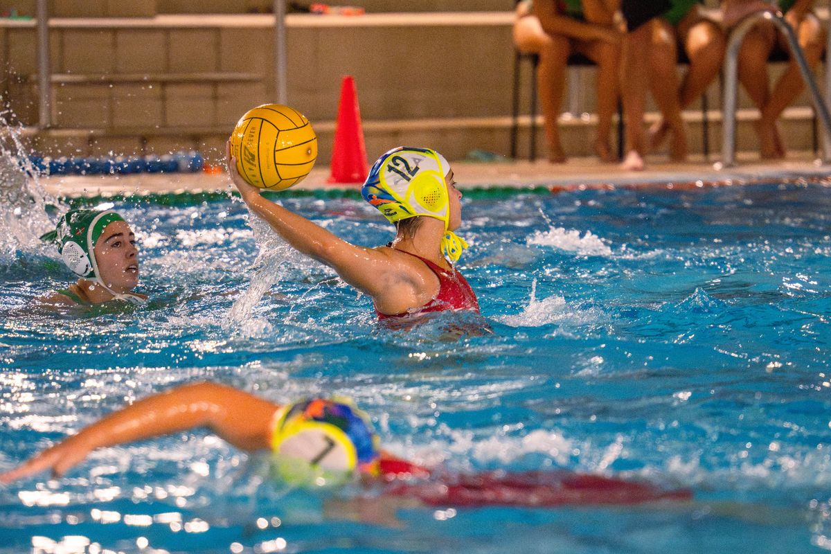 Jugadora de waterpolo con gorro amarillo avanza con el balón mientras una rival se acerca para defender.